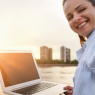 Eine Frau sitzt am Ufer, hat ihren Laptop aufgeklappt auf ihrem Schoß und macht ein Selfie im Sonnenuntergang