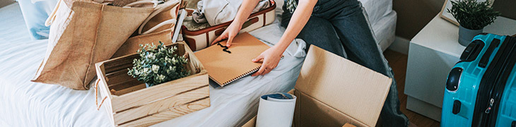 Jemand sitzt auf einem vollgräumten Bett, rundherum Kisten und Koffer, in denen eingepackt wird.