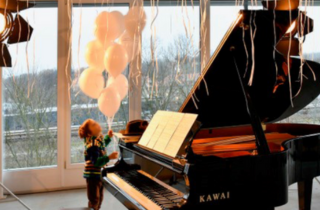 Kind steht neben einem Klavier und hält weiße Luftballons in der Hand