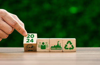 Eine Hand dreht einen Würfel um, auf welchem 2024 und 2024 steht. Daneben sind weitere Würfel, die Symbolbilder zum Thema Nachhaltigkeit enthalten.