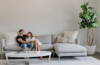 Ein Paar sitzt im Wohnzimmer auf einer hellen Couch und lächelt sich an.